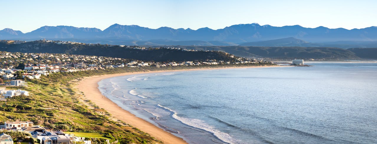 Curved sandy beach with waves, coastal houses, and mountains under clear blue sky.