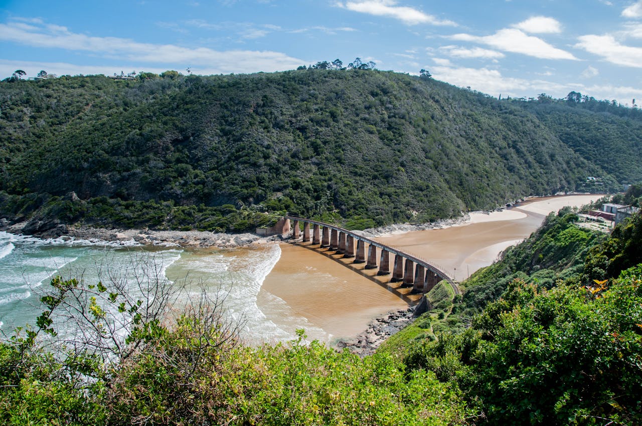 Curved bridge spanning a sandy river mouth with green hills and blue sky in the background.