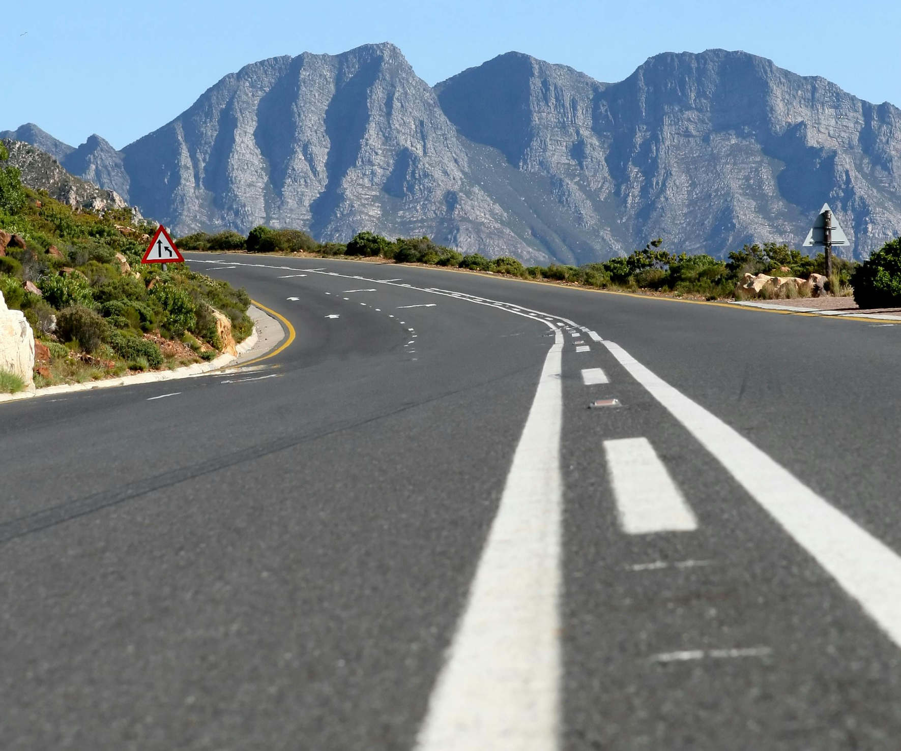 Empty paved road curving left with traffic signs and rocky mountains under a clear blue sky.