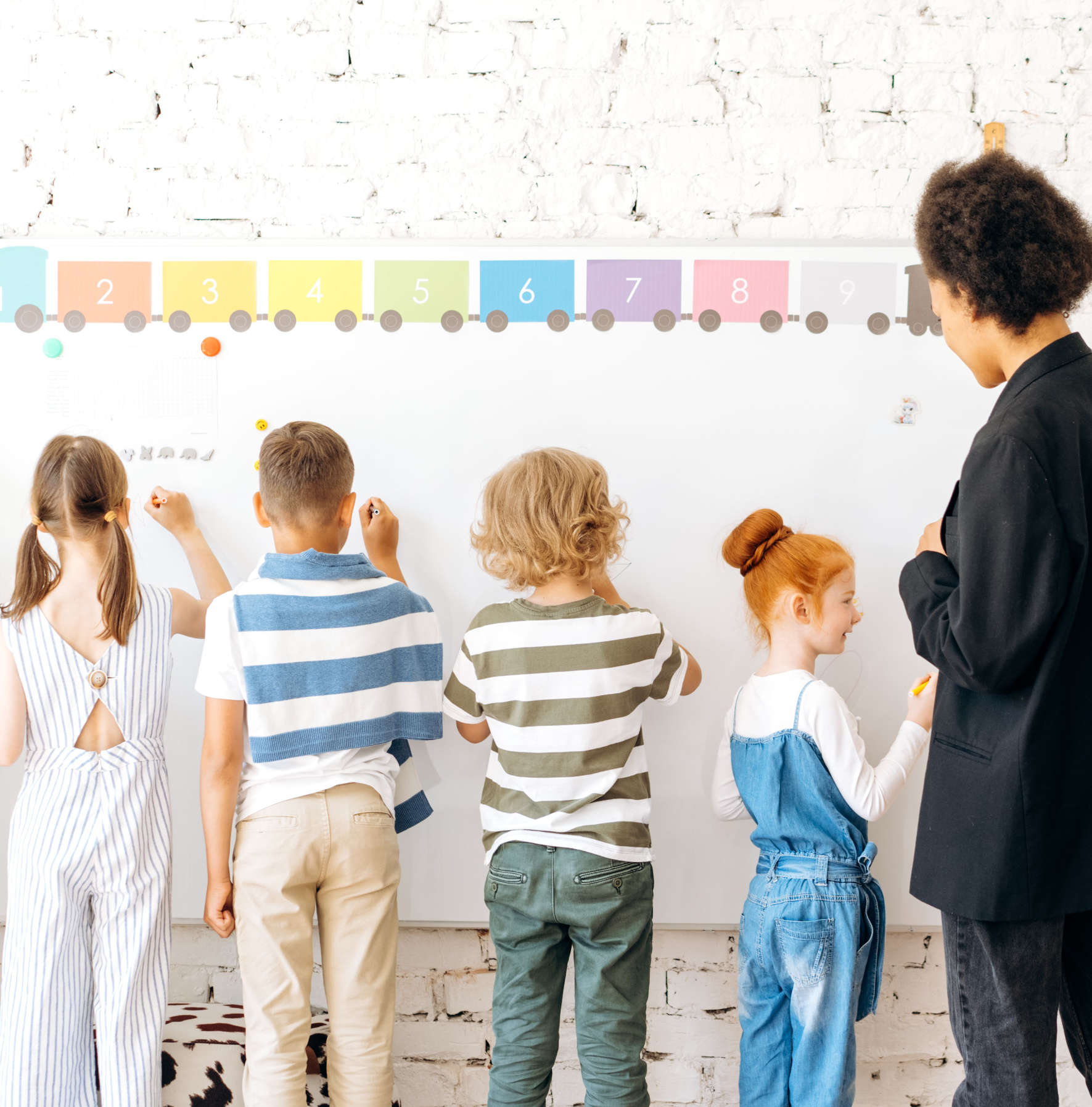 Four children writing on a whiteboard with a teacher watching in a classroom with a number train above.