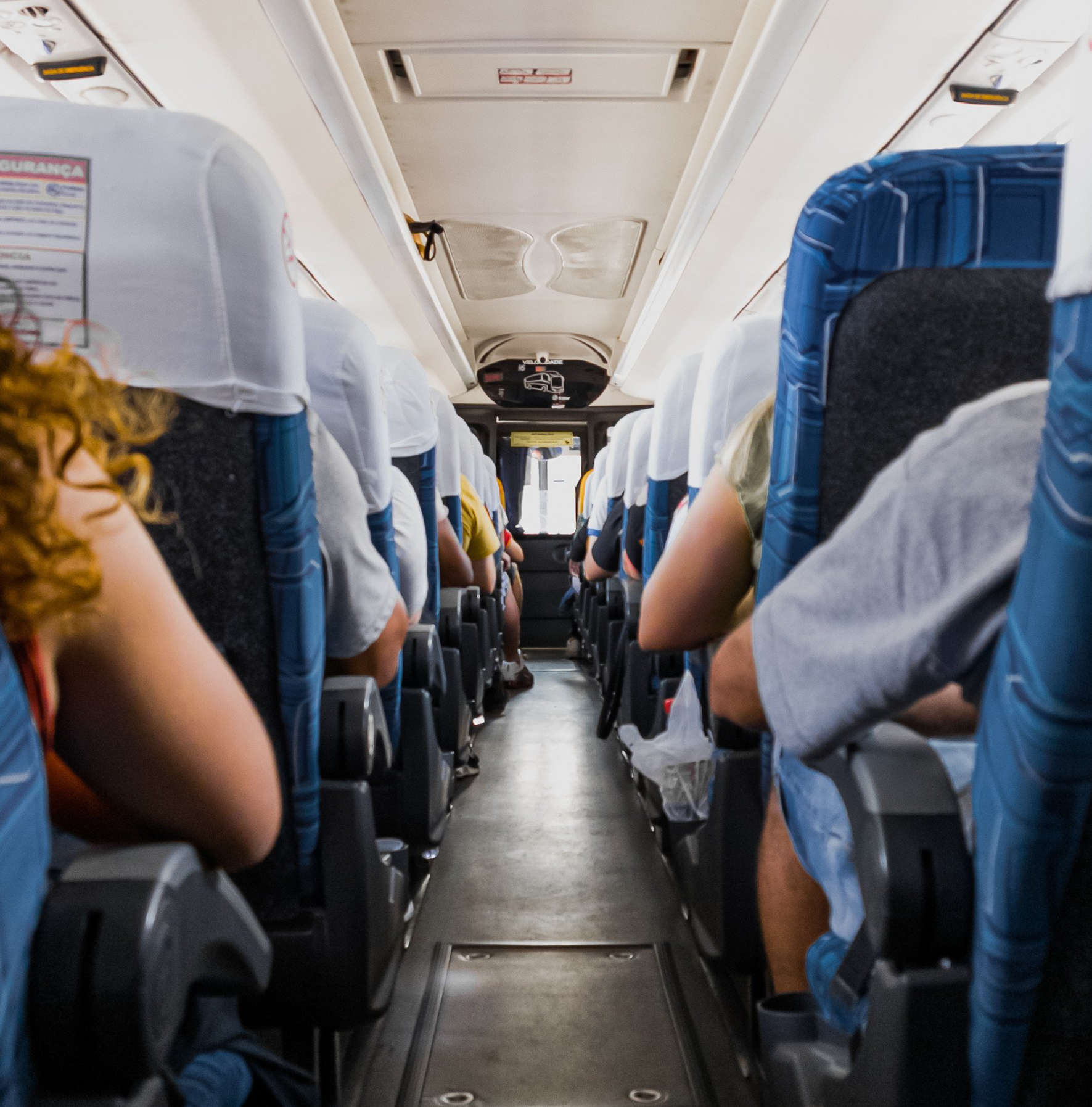 Passengers seated inside a bus with blue seats and white headrest covers, viewed from the aisle.