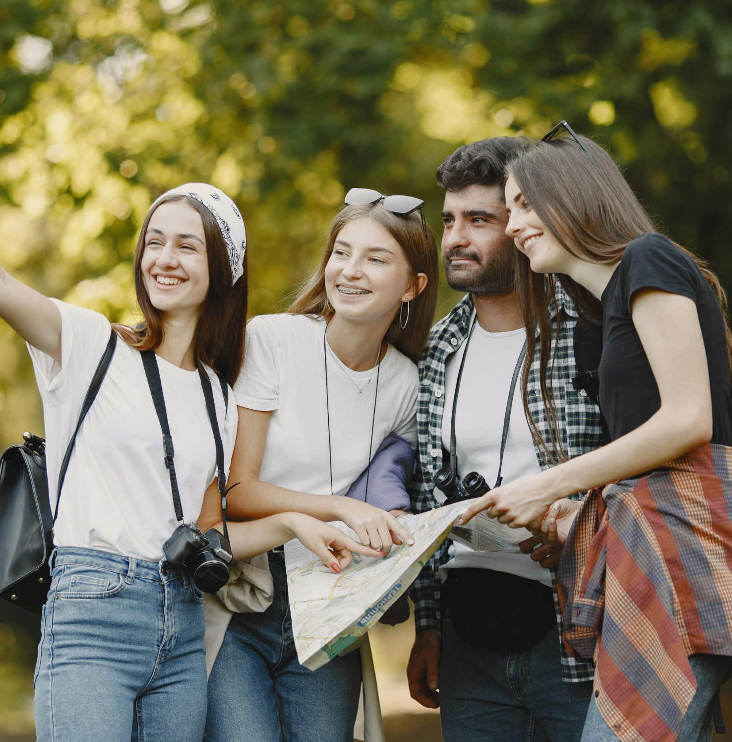 Group of four young adults outdoors looking at a map and taking a selfie, smiling amid green trees.